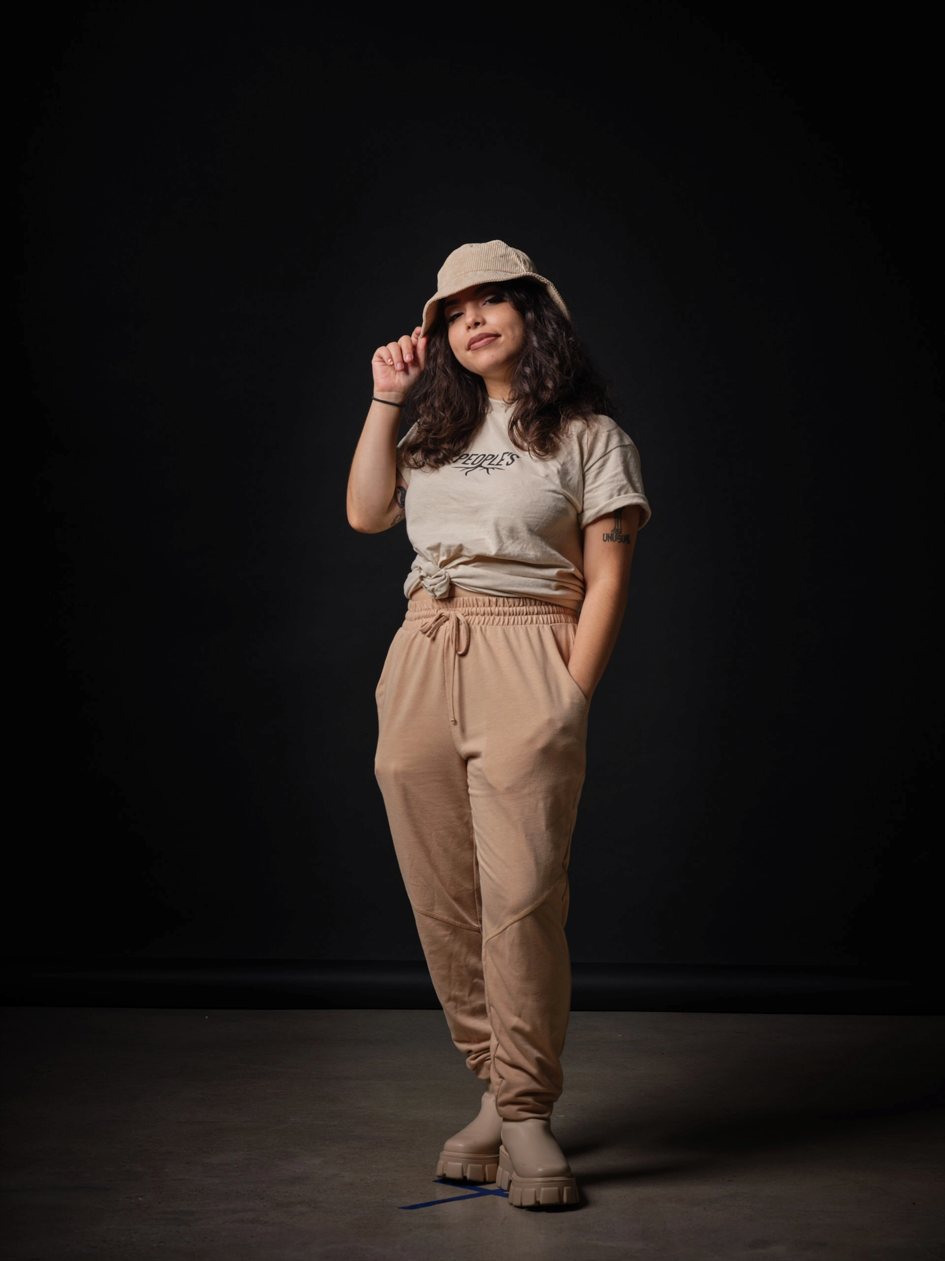Full-body studio portrait by San Francisco photographer; featuring a woman in tan streetwear and a bucket hat, captured on GFX medium format with a single Broncolor Para 133 for a moody, high-end aesthetic.